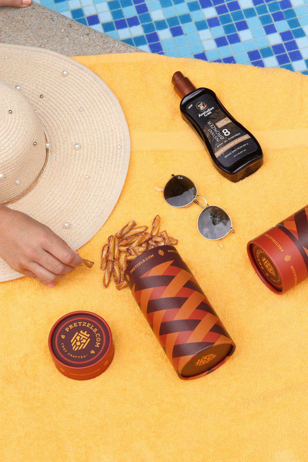 Yellow towel with pretzels, sunscreen, sunglasses, and a hat by a pool.