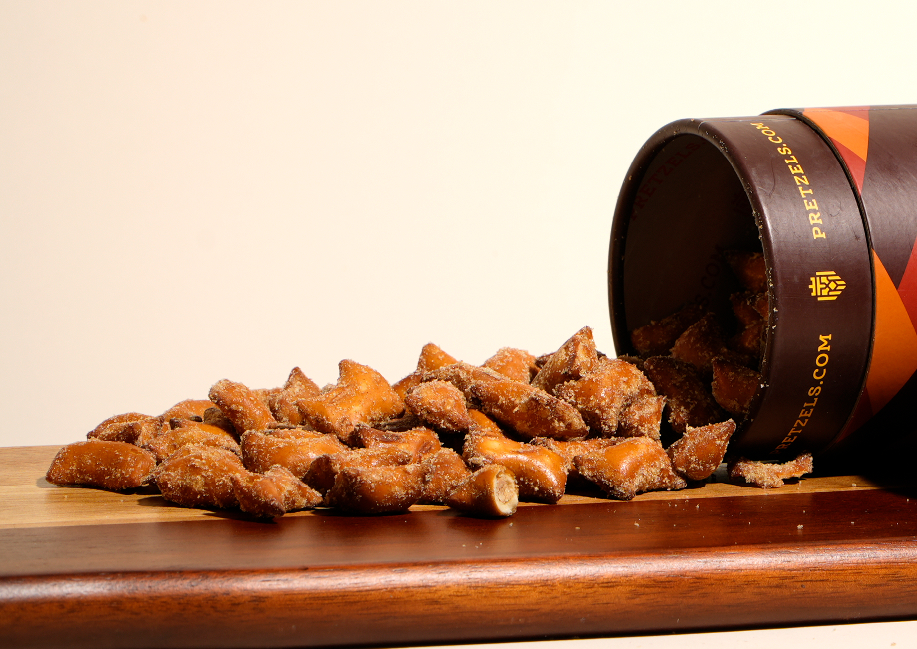 Seasoned pretzel pieces spilling out of a cylindrical container onto a wooden board against a white background.