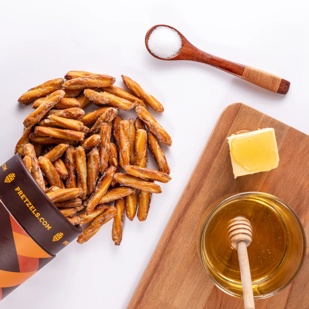 Pretzels, honey in a bowl, butter, and a spoon of salt on a wooden board.