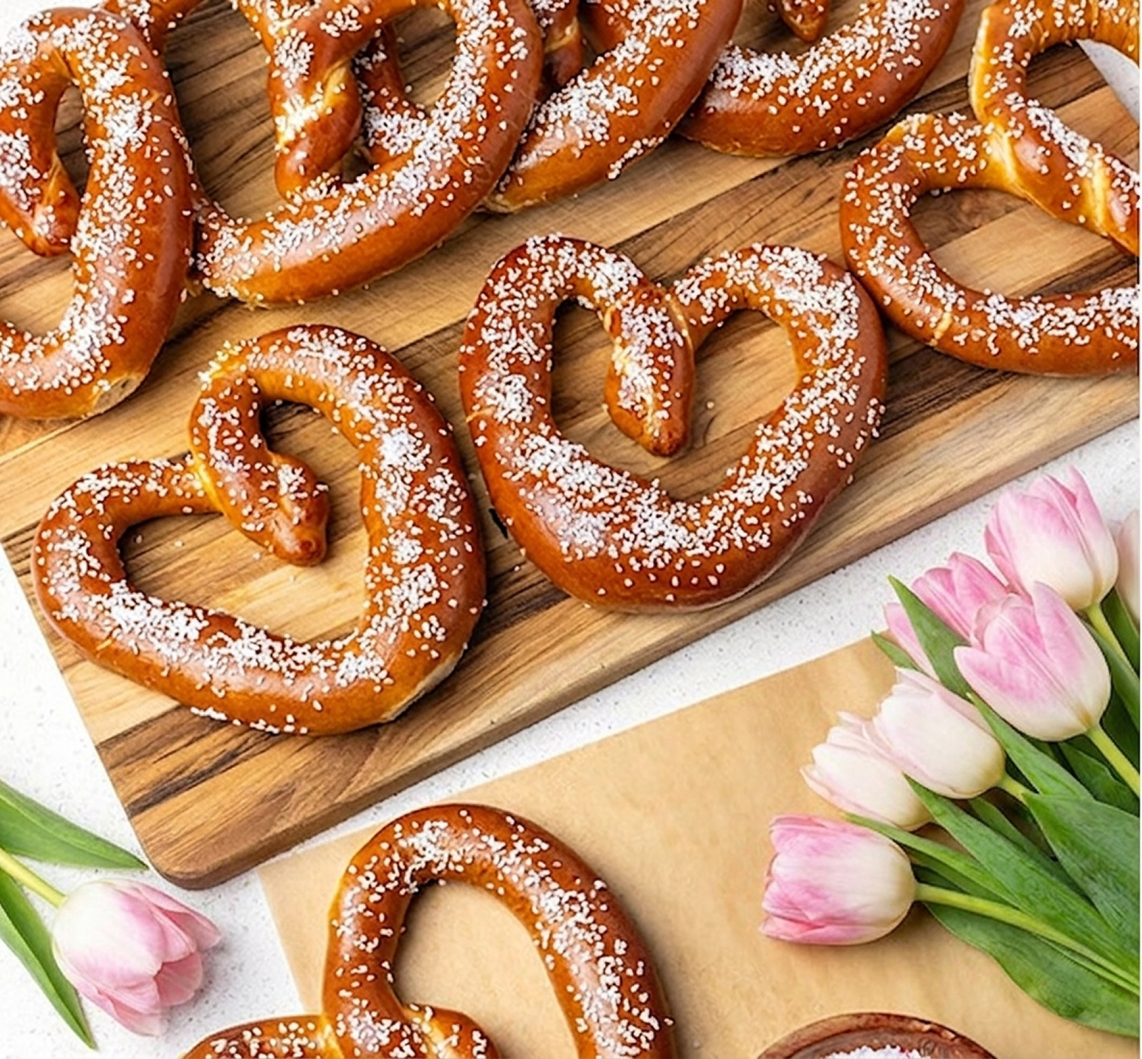An overhead view of heart-shaped soft pretzels sprinkled with salt, arranged with fresh pink tulips on a table.