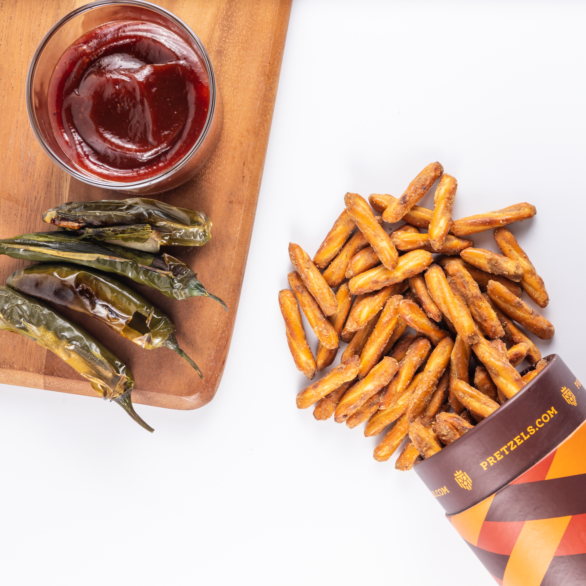 An overhead shot of pretzel sticks spilling from a cup, next to roasted peppers and dipping sauce.