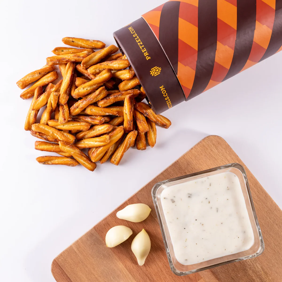 Container spilling pretzel sticks next to garlic cloves and a bowl of white sauce.
