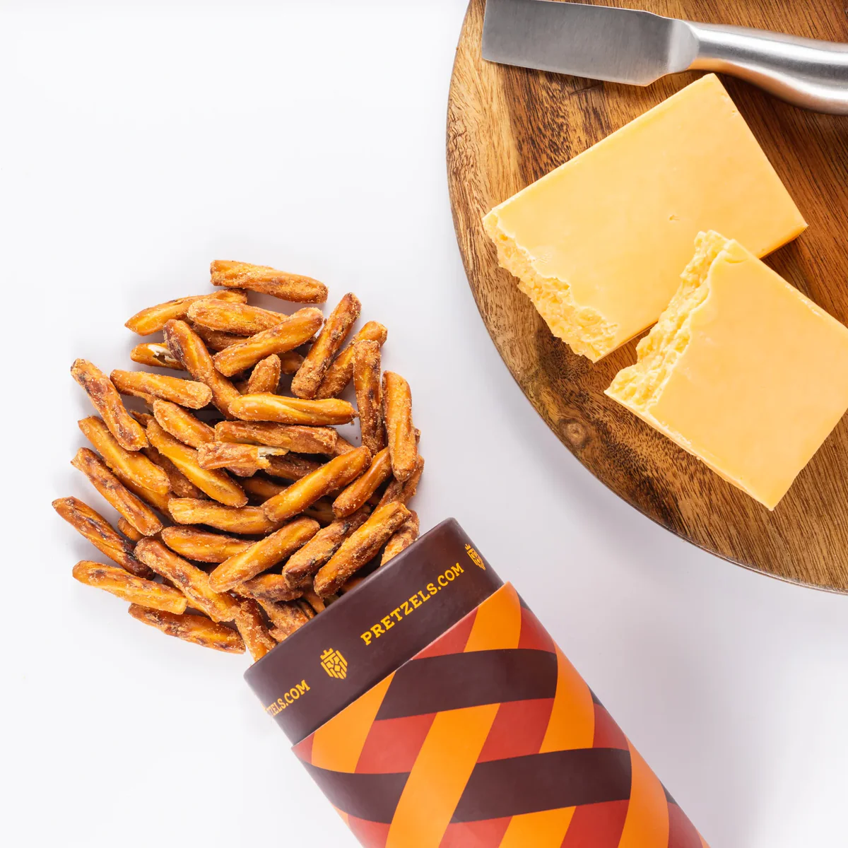 Pretzel sticks spilling from a container near cheese blocks on a wooden board.