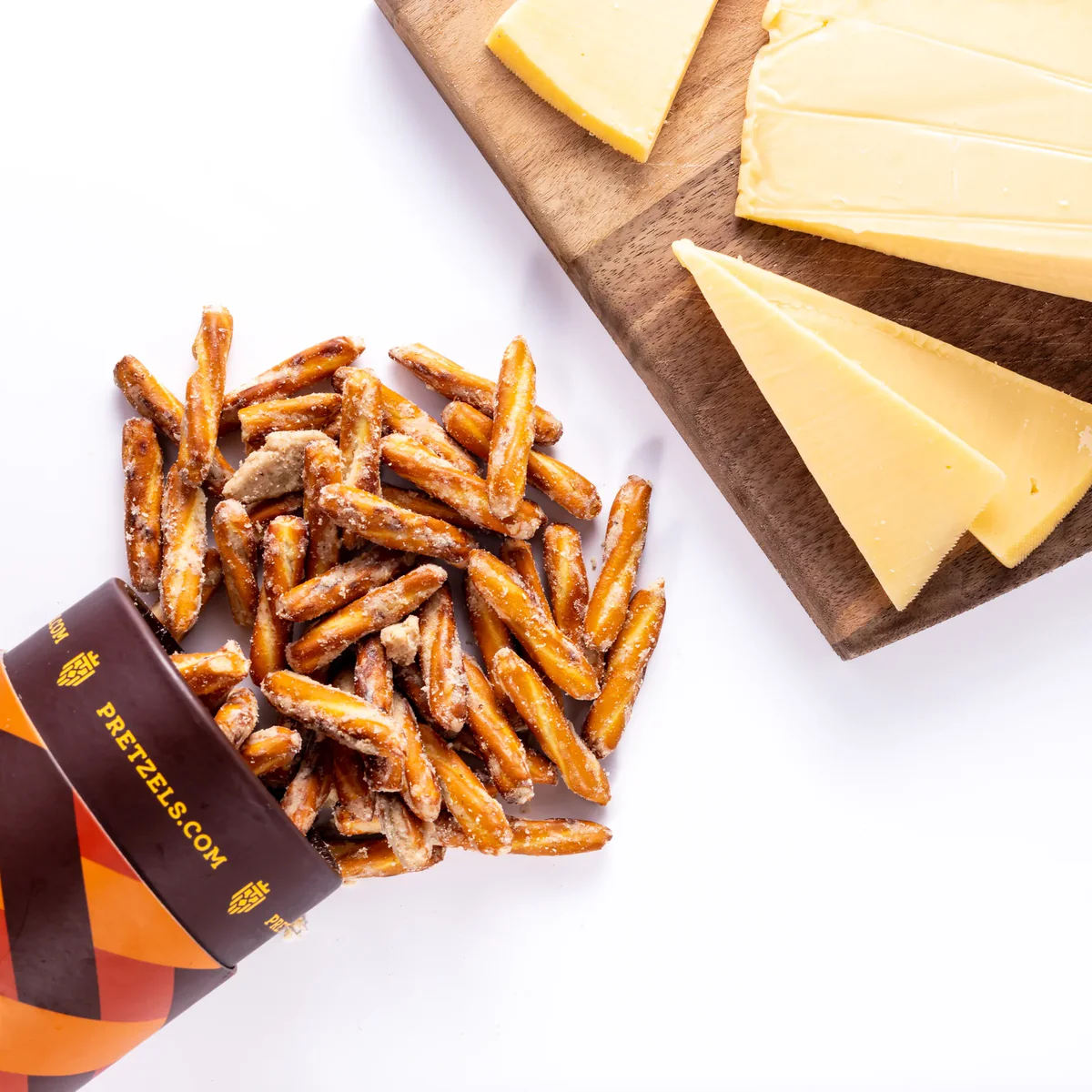 Pretzel sticks spill from a container next to sliced cheese on a wooden board.
