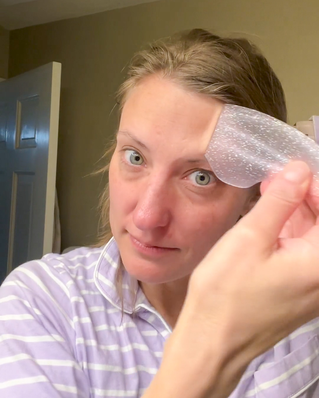A woman in striped pajamas applies a clear, shimmery patch to her forehead while looking at the camera.