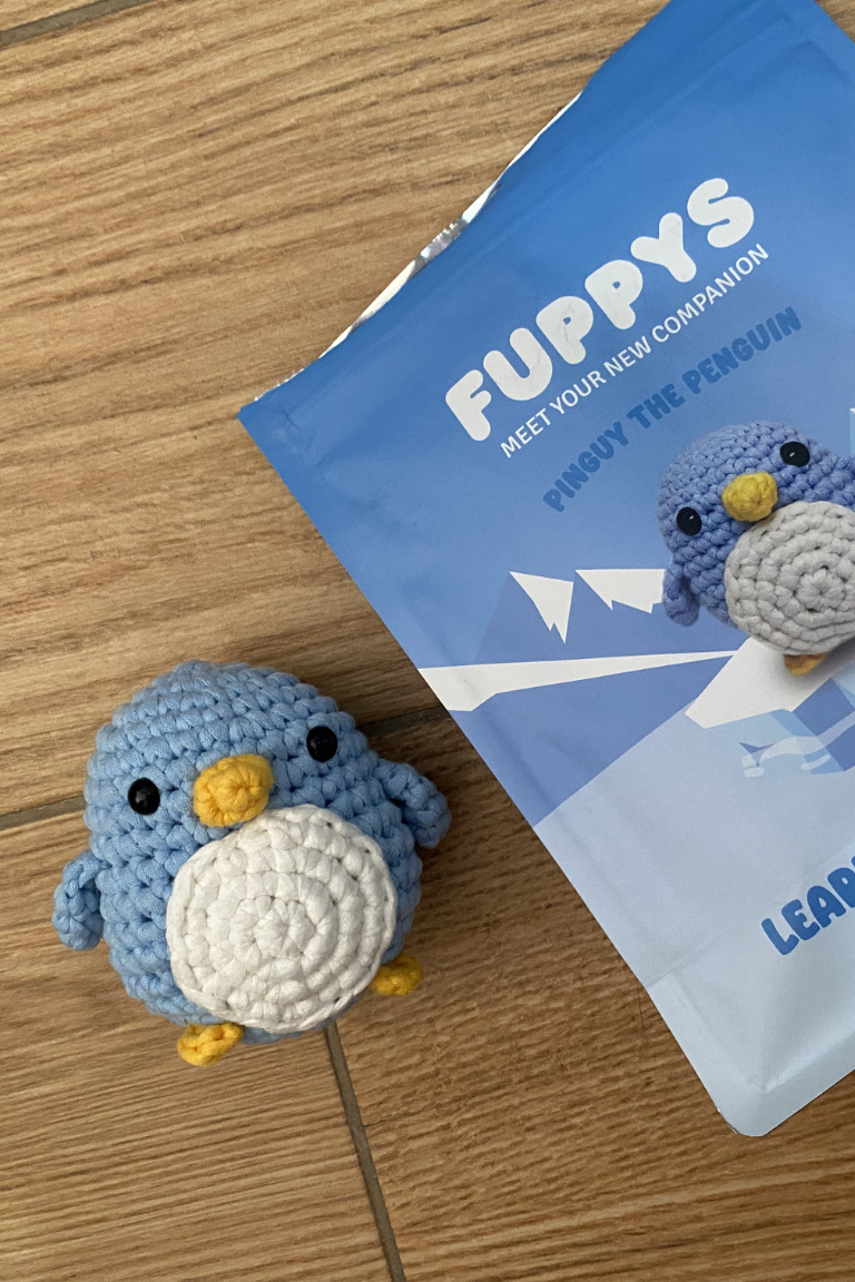 A blue crocheted penguin toy sits next to its blue packaging on a wooden surface.