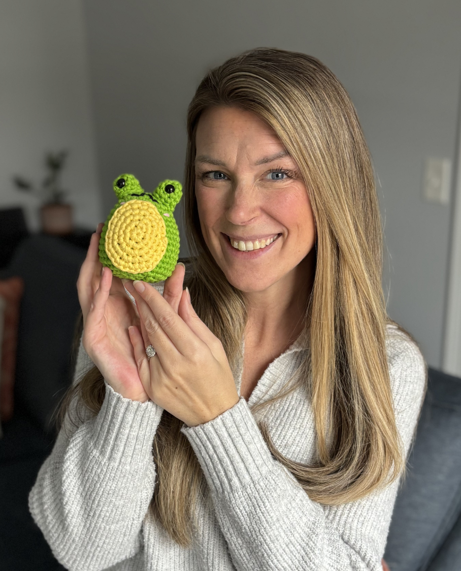 A smiling woman in a grey sweater holds up a small, green and yellow crocheted frog.
