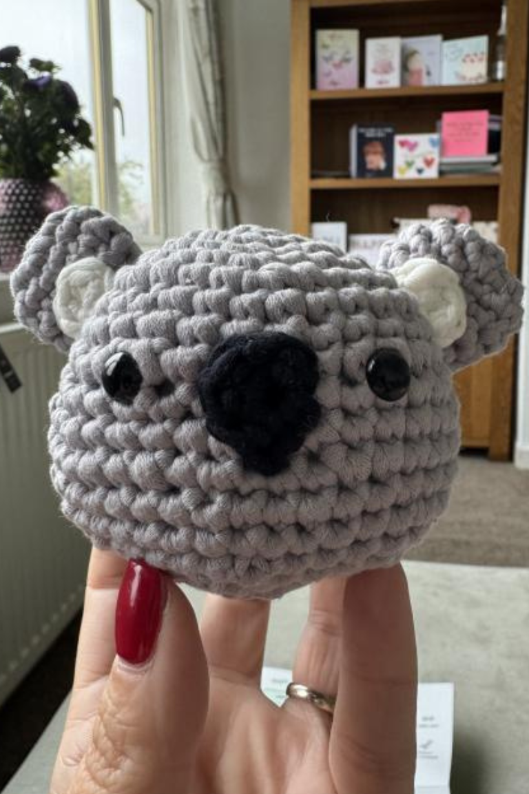 A hand with red nail polish holding up a small, gray crocheted koala head.