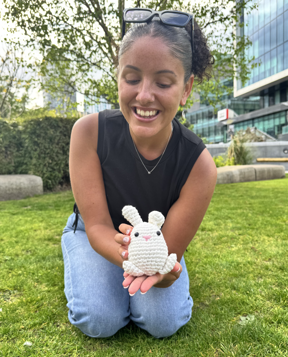 A woman smiles while kneeling on grass, holding a small white crocheted bunny in her hands.