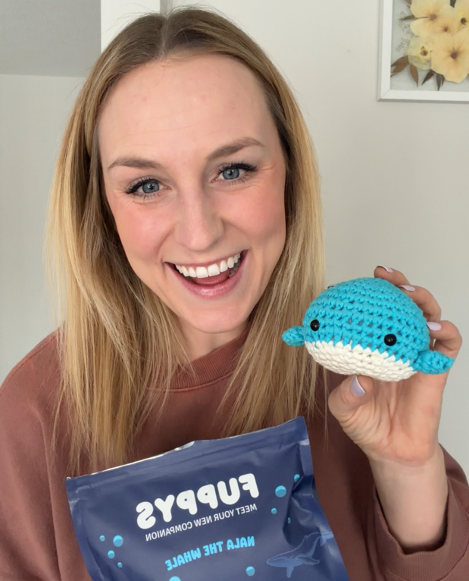 A smiling woman holds a blue crocheted whale toy and a Fuppys product bag.