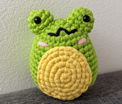 A close-up of a green crocheted frog toy with a round yellow belly.