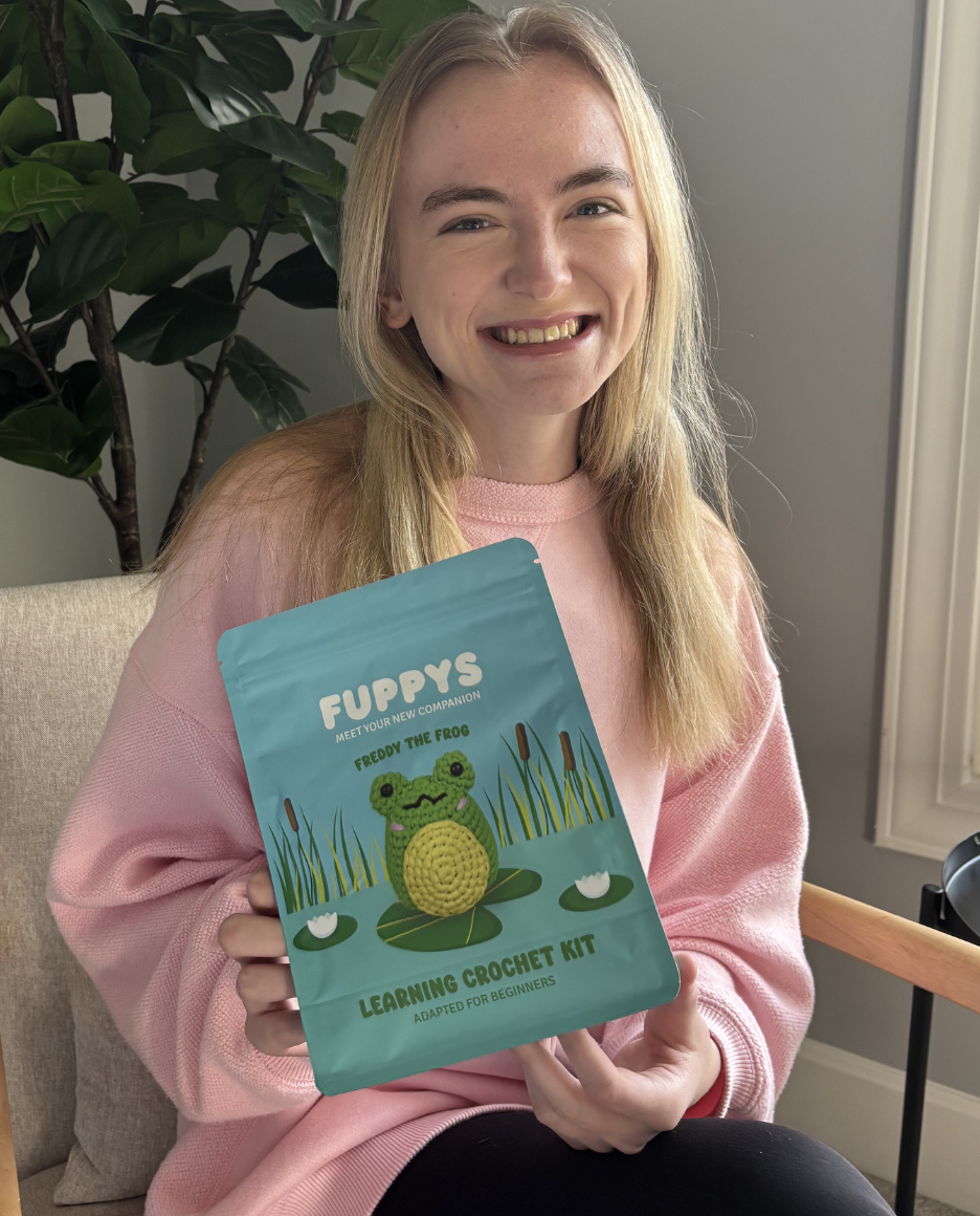 A smiling woman in a pink sweater holds up a learning crochet kit for a frog.