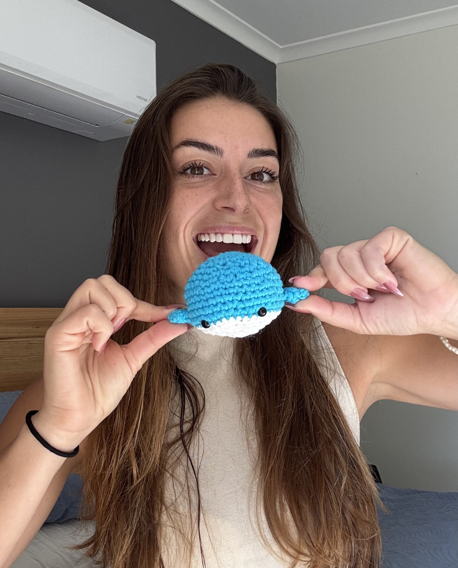 A smiling woman holds a small blue and white crocheted whale toy in front of her mouth.