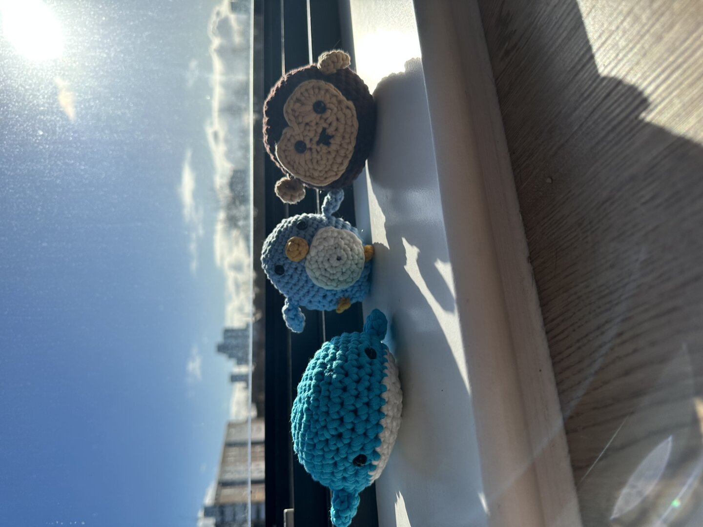 Three small crocheted animals, a whale, a penguin, and a monkey, sit on a sunny windowsill.