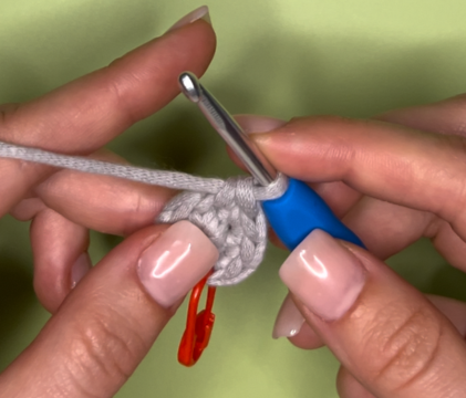 A close-up of hands crocheting a small, gray circle with a blue-handled hook and a red stitch marker.