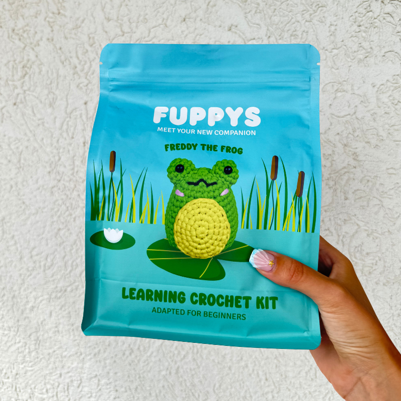 A blue crochet kit package featuring a frog design held by a hand.