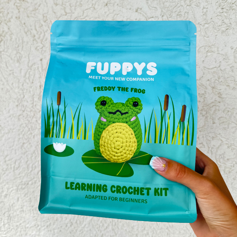 A hand holding a blue pouch for a 'Freddy the Frog' learning crochet kit for beginners.