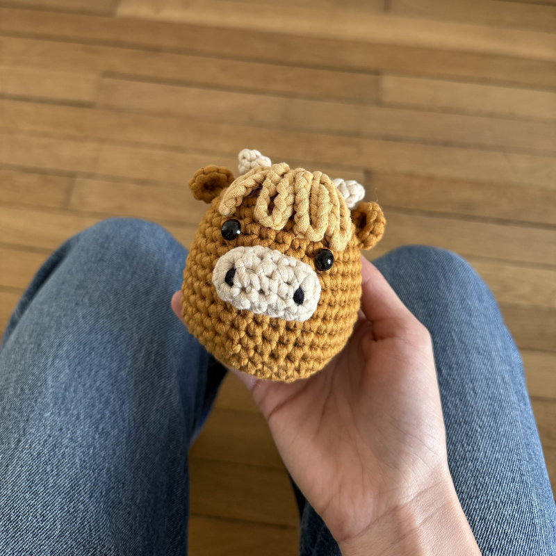 A person's hand holding a small, crocheted brown cow head with a cream-colored snout.