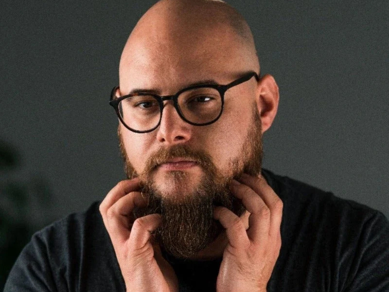 A bald man with a beard and glasses touches his beard with both hands against a grey background.