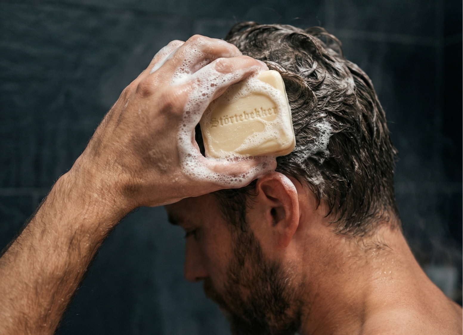 A man lathers his wet hair with a bar of soap against a dark background.