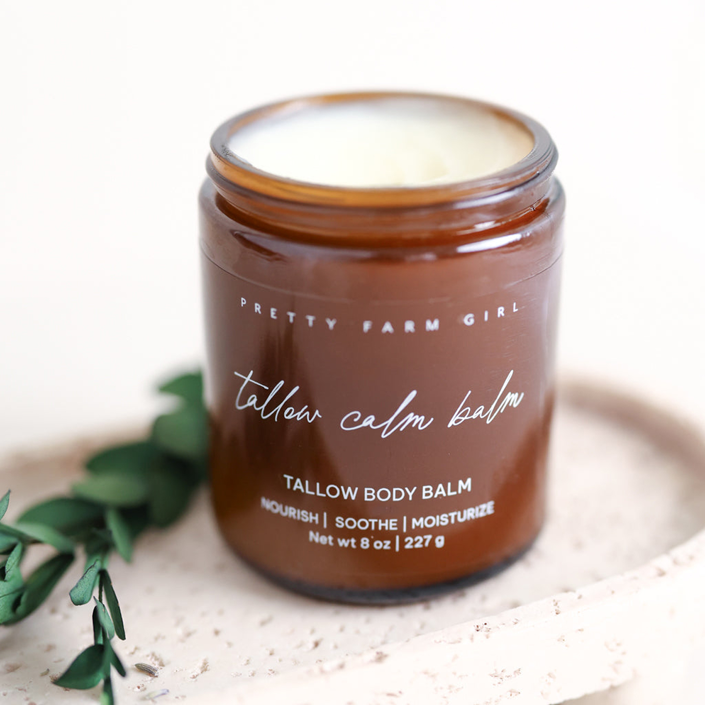 An open amber glass jar of Pretty Farm Girl Tallow Calm Balm sitting on a textured tray with greenery.
