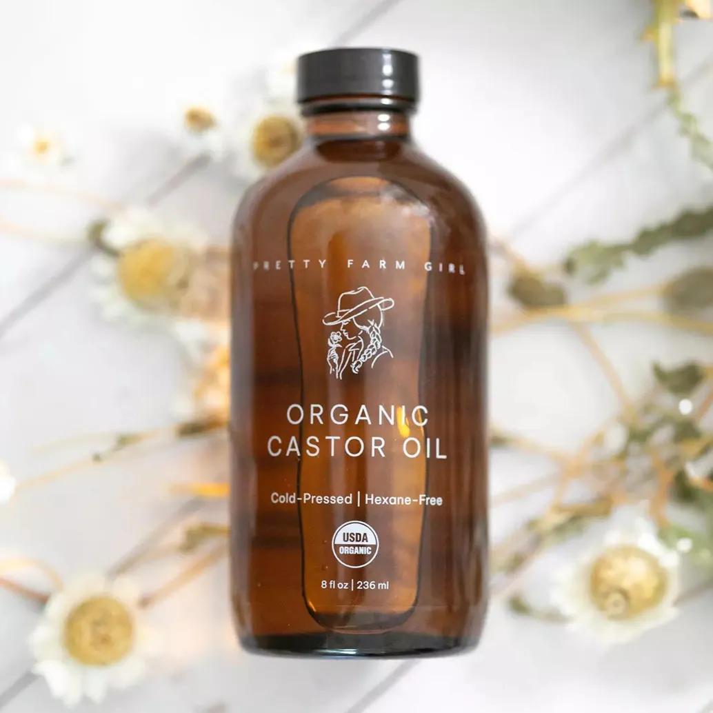 A brown bottle of Pretty Farm Girl Organic Castor Oil sits on a white surface with dried flowers.