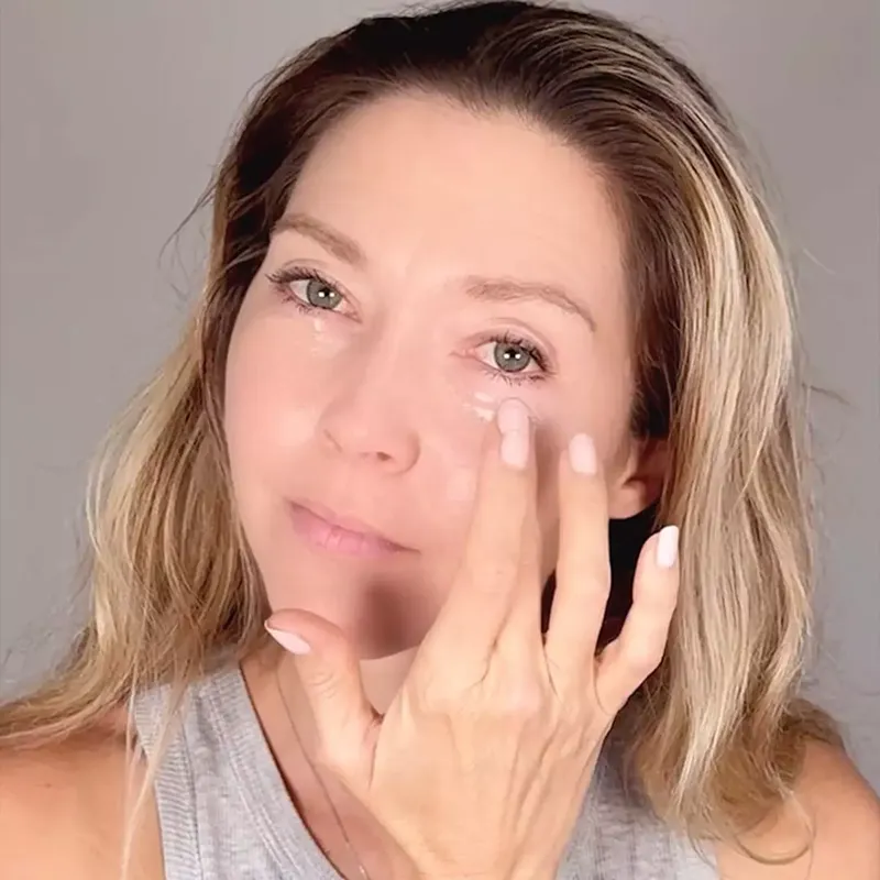 A woman with blonde hair and green eyes applying a white cream under her right eye.