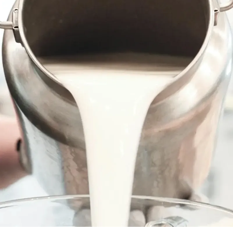 A thick, creamy white liquid being poured from a large stainless steel pot into a bowl below.
