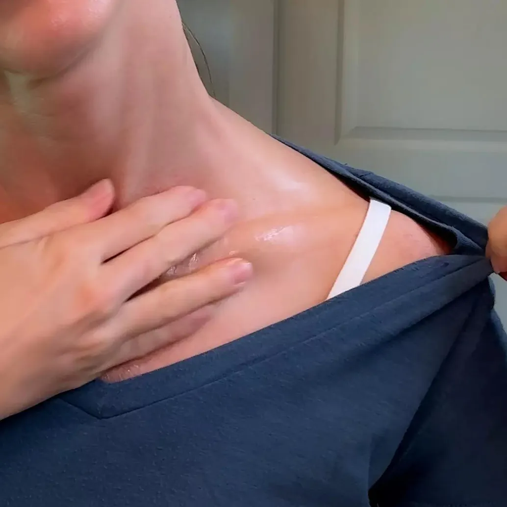 A close-up of a person applying a shiny lotion to their neck and collarbone.