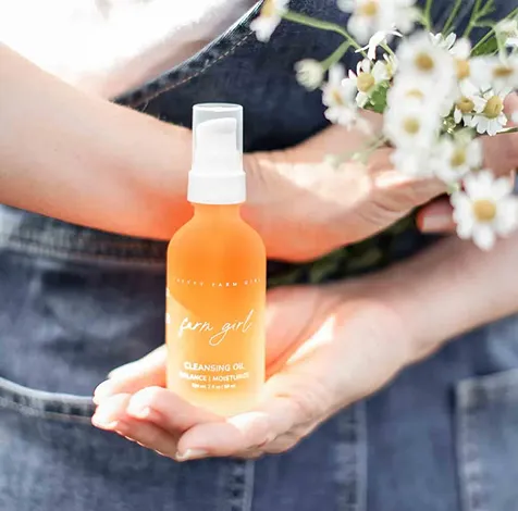 A person holds a bottle of Farm Girl Cleansing Oil and a small bouquet of white flowers.