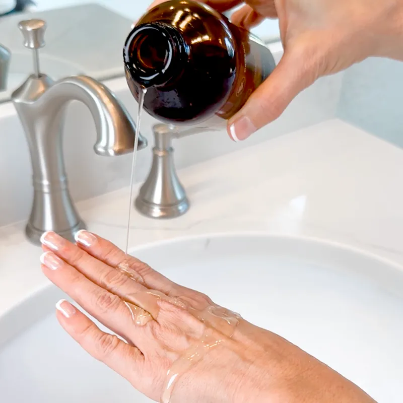 A person pours a clear liquid from an amber glass bottle onto their hand over a sink.