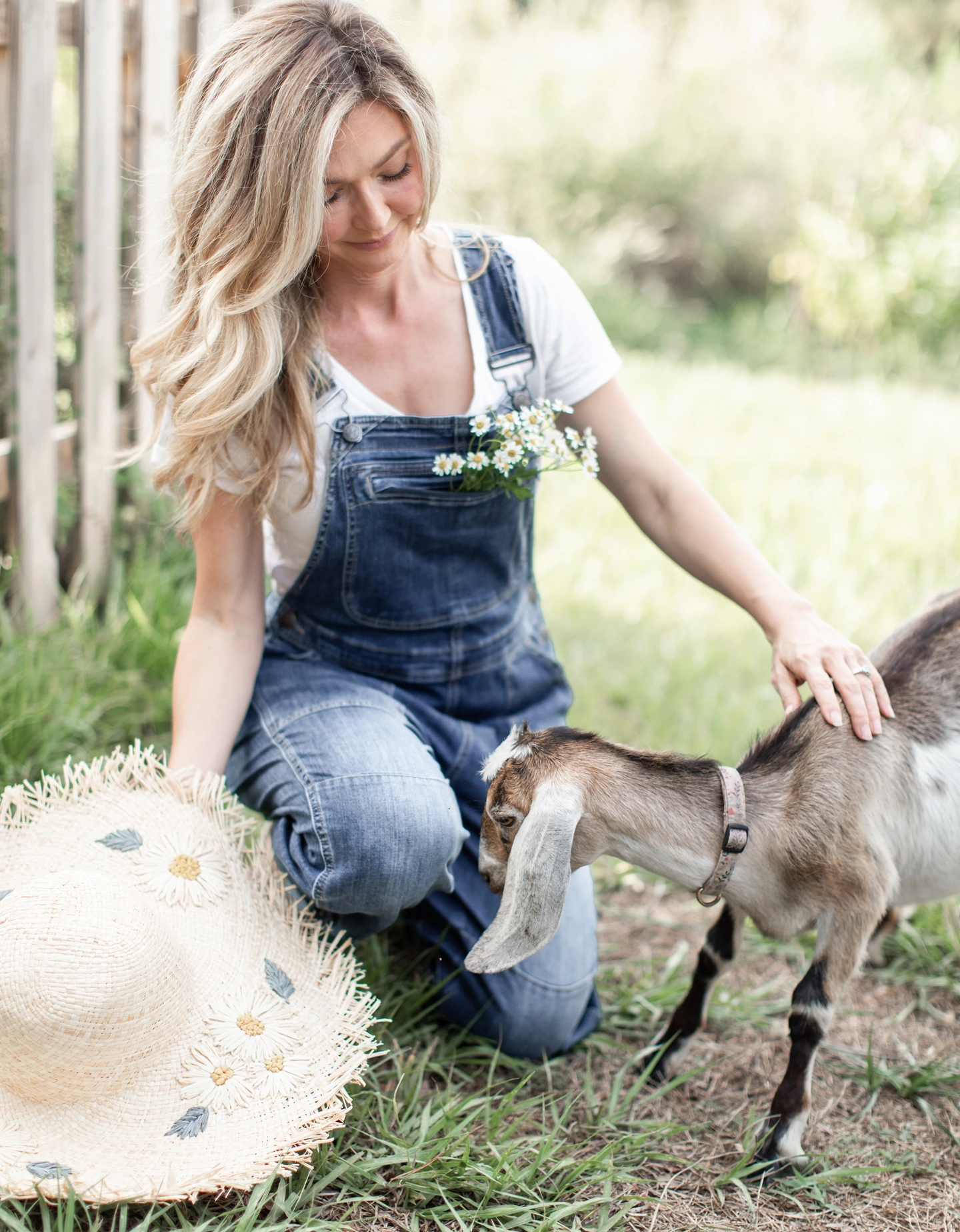 A woman in denim overalls with daisies in her pocket kneels in the grass and pets a goat.
