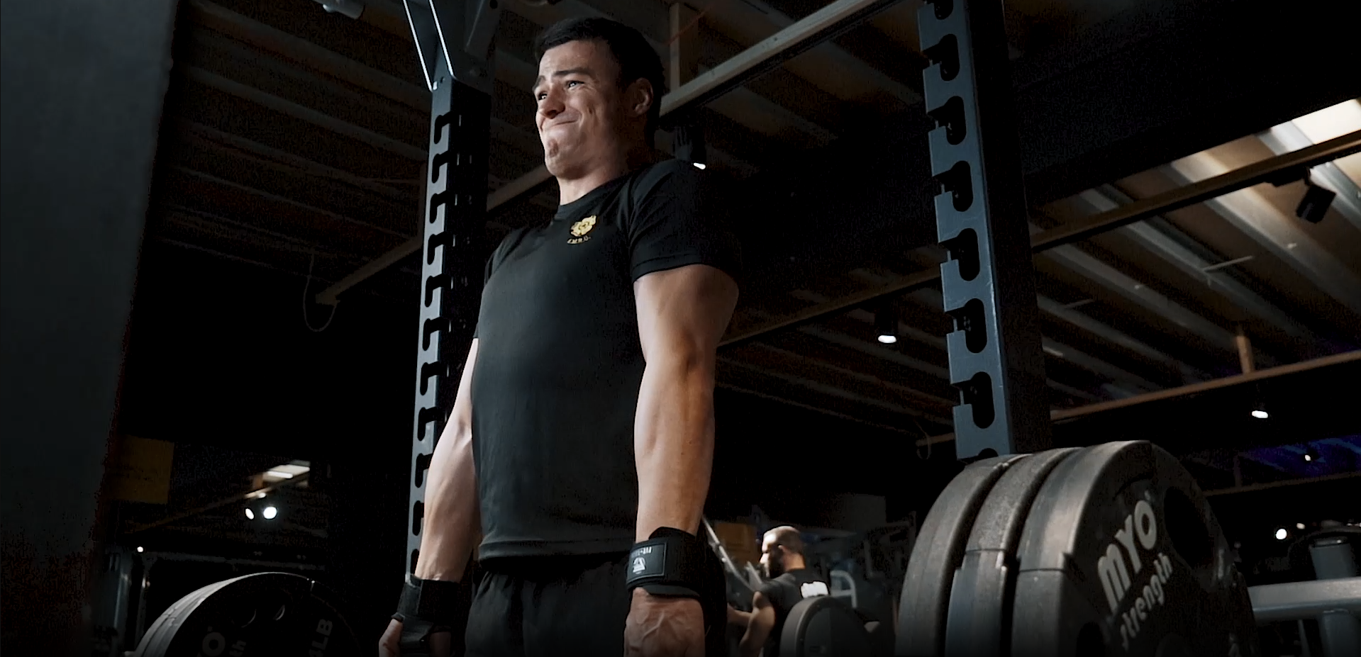 Person lifting weights in a gym, wearing a black shirt and wrist straps.