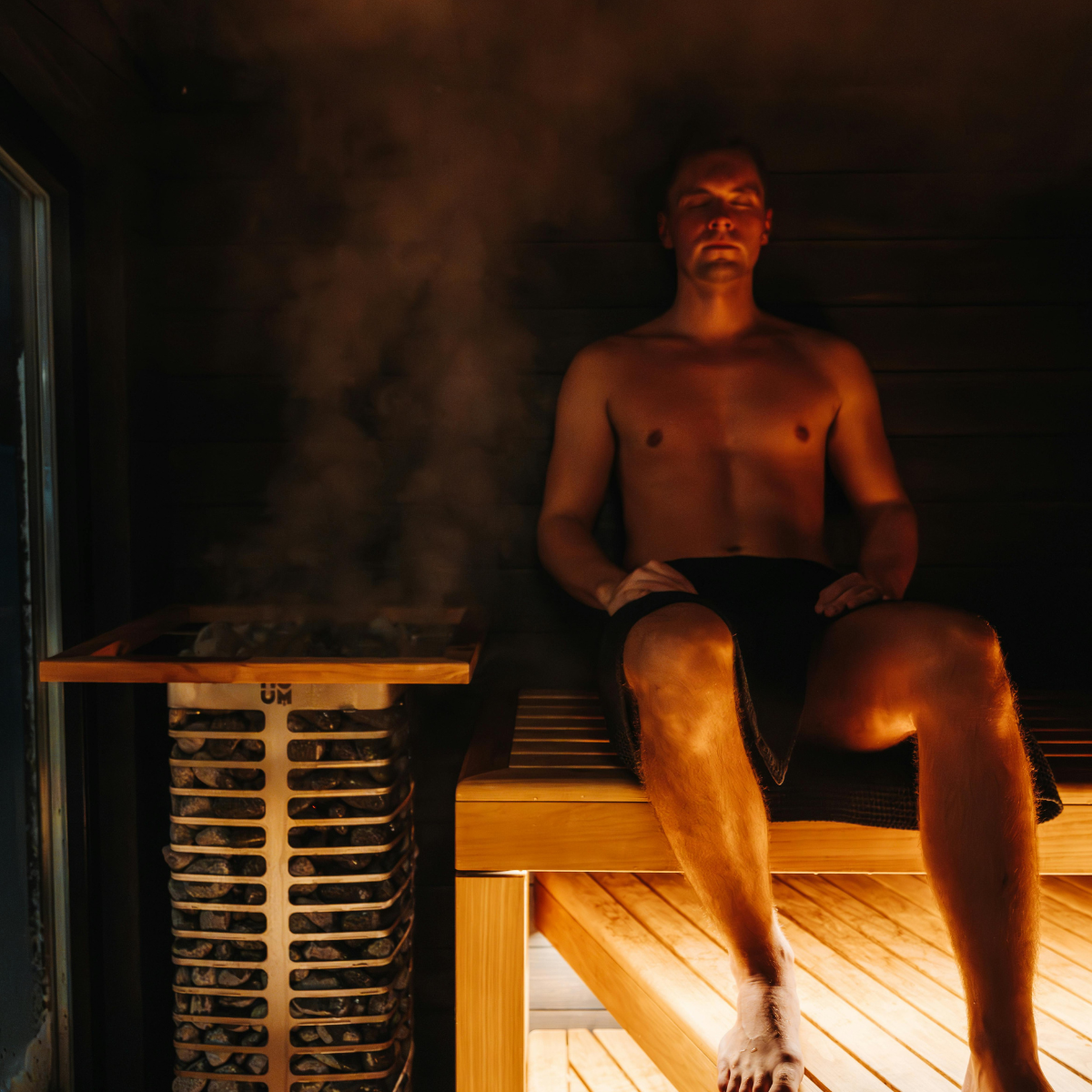 A shirtless man sits on a wooden bench inside a steamy sauna next to a rock heater.