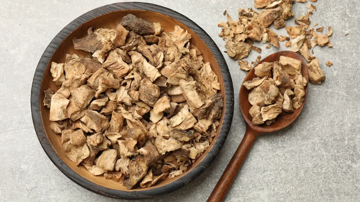 An overhead shot of dried, chopped burdock root in a wooden bowl and spoon on a grey surface.