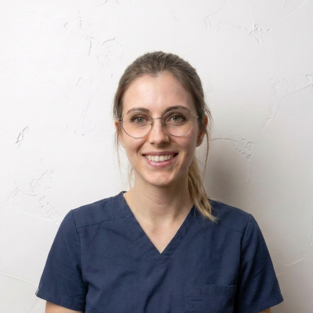A smiling woman with glasses and light brown hair tied back, wearing blue scrubs against a white wall.
