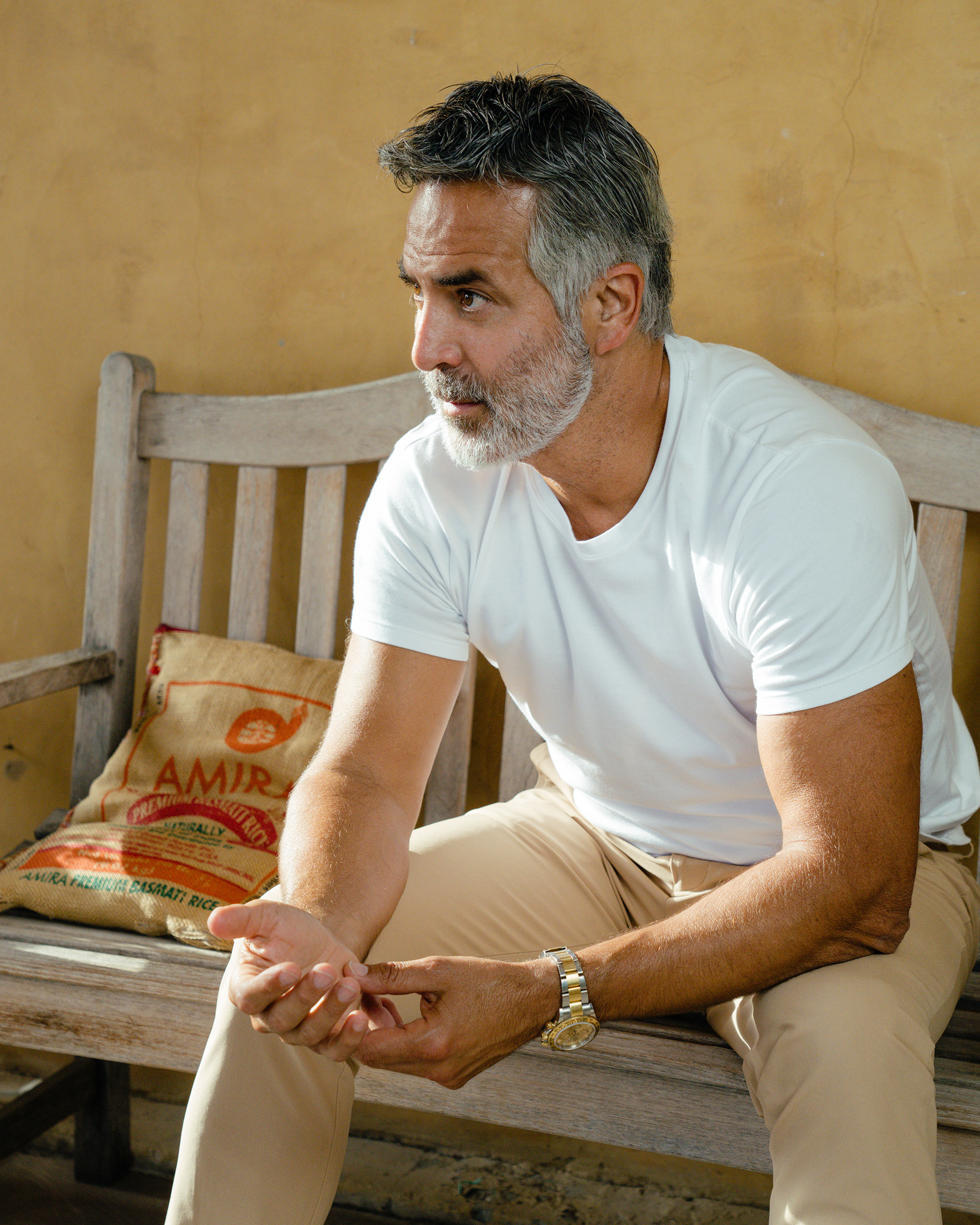 Man with gray hair and beard sitting on a wooden bench indoors.