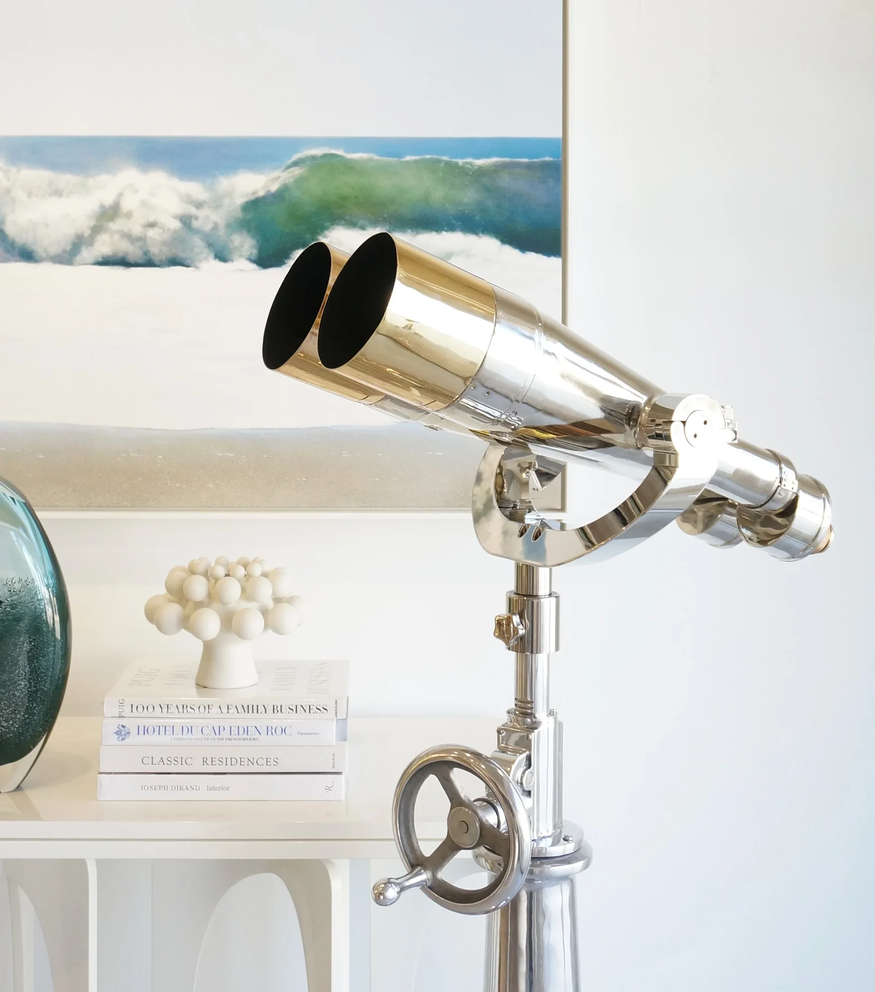 A large chrome binocular telescope on a stand, with a painting of an ocean wave in the background.