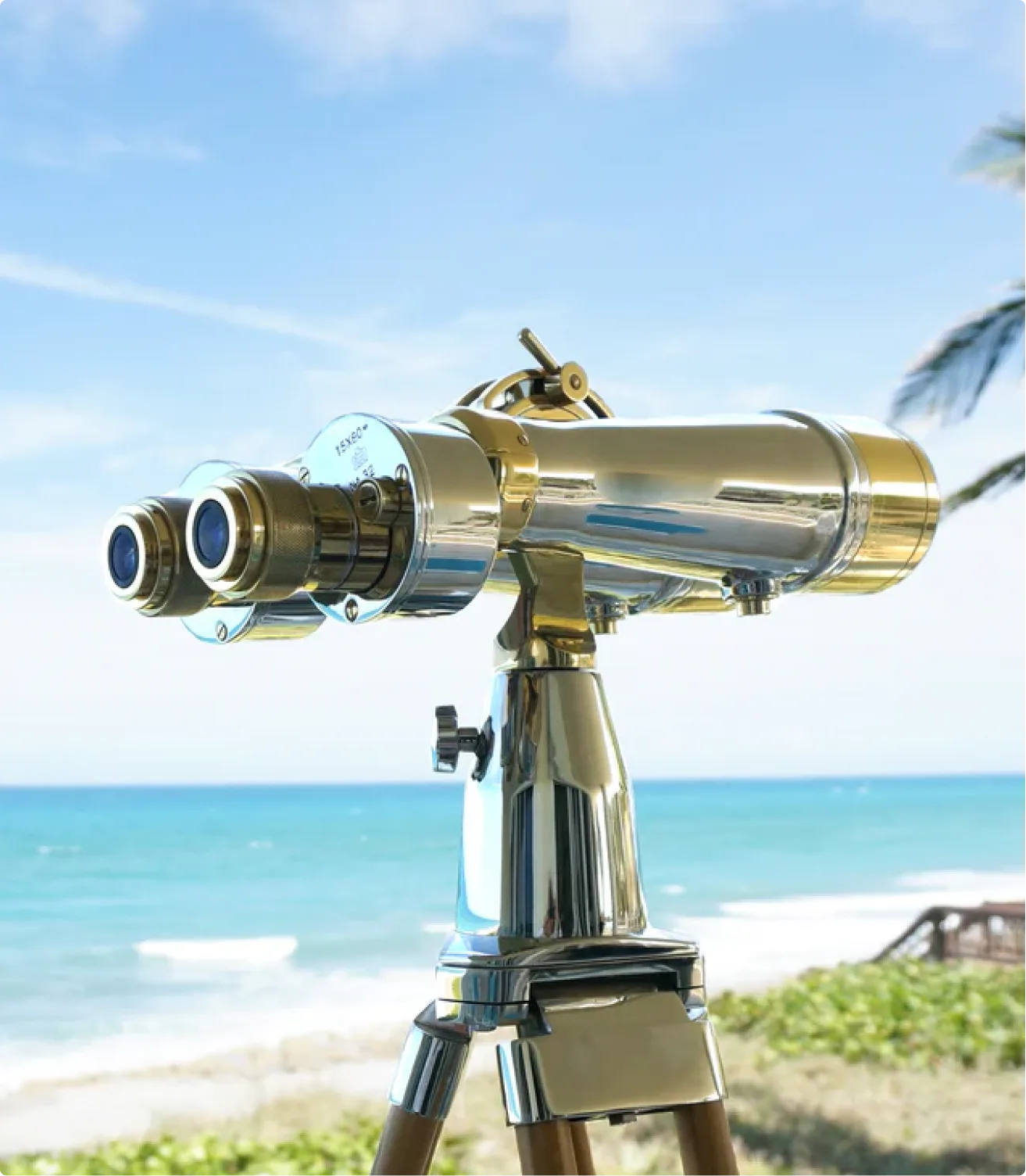 Large, metallic binoculars on a tripod stand with a beach and ocean view in the background.