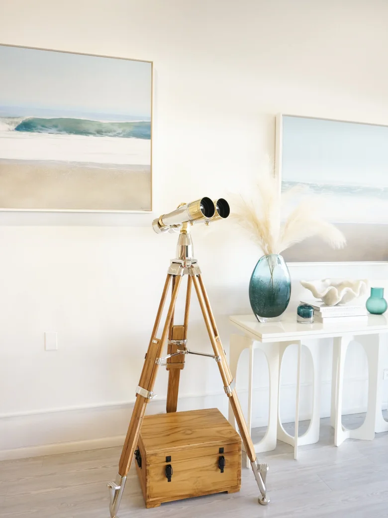 A pair of binoculars on a wooden and chrome tripod in a coastal-themed room with beach paintings.