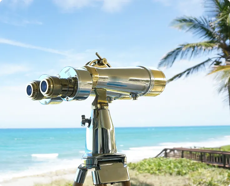 A close-up of a pair of large, chrome binoculars on a tripod overlooking a sunny beach and ocean.
