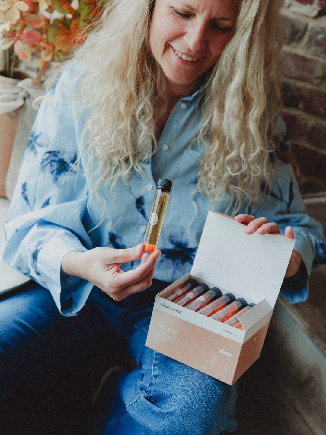 A smiling woman with long blonde hair holds up a small vial from a box of supplements.
