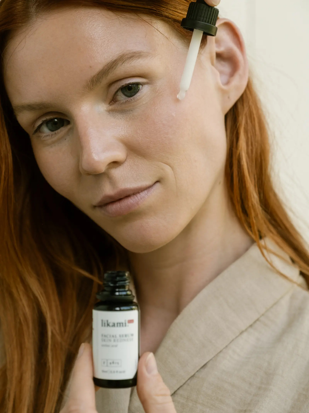 A woman with red hair applies facial serum to her cheek with a dropper while holding the bottle.