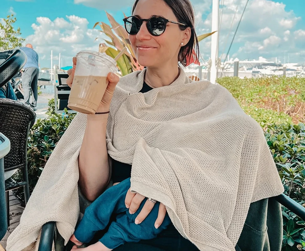A woman in sunglasses holds an iced coffee while a baby sits on her lap under a cover.