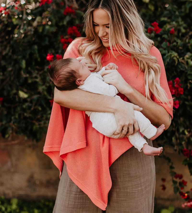 A woman with long blonde hair smiles while holding a newborn baby in front of a red-flowered bush.