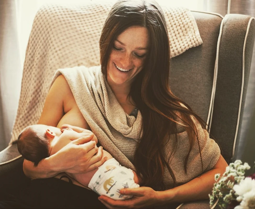 A smiling woman with long brown hair breastfeeds her newborn baby while sitting in an armchair.