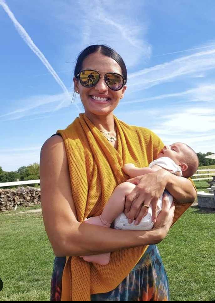 Woman smiling and holding a baby outdoors on a sunny day.
