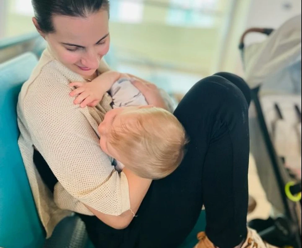 A woman with dark hair pulled back smiles as she breastfeeds a blond baby in her lap.