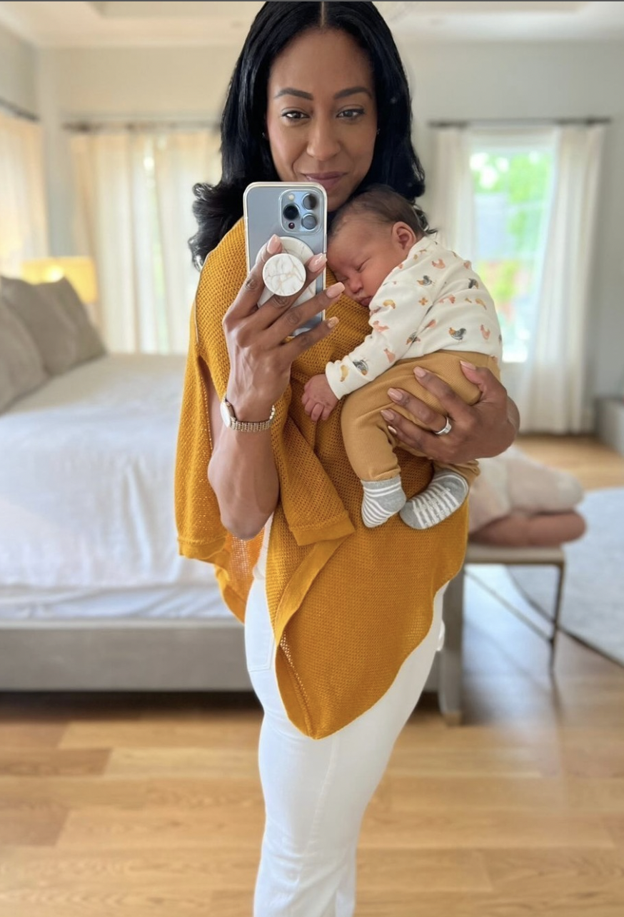 Woman holding a sleeping baby, taking a mirror selfie in a bedroom.