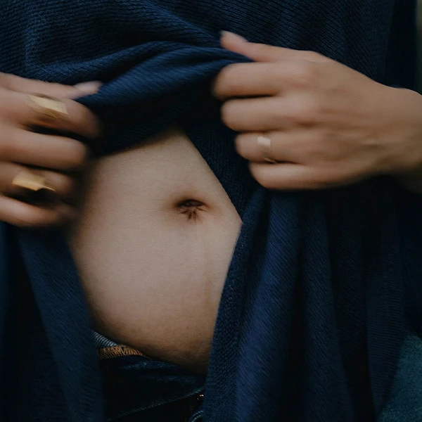A person's hands, adorned with rings, lift a dark blue sweater to expose their navel.